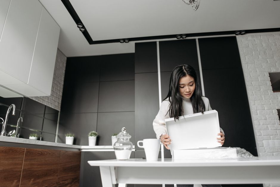 Woman enthusiastically unboxing a package in a modern kitchen setting, showcasing online shopping experience.